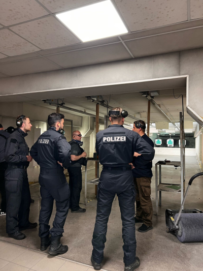 Policijas darbinieki šaušanas apmācībās slēgtā šautuvē — instruktors sniedz norādījumus pie šaušanas stenda.