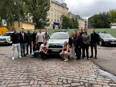 Grupas foto pilsētvidē pie Latvijas Valsts policijas patruļautomašīnas — civilā apģērbā tērpti jaunieši pozē kopā uz bruģētas ielas.