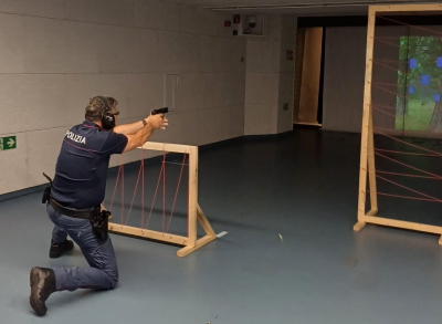 Policists ar uzrakstu 'Polizia' uz krekla trenējas šaušanā telpās, nometies uz viena ceļgala un tēmējot ar pistoli caur šķēršļu rāmi ar saspriegtām auklām. Fonā redzams digitālais mērķis uz sienas.