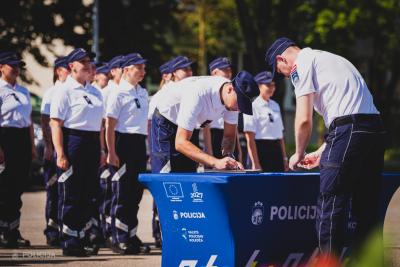 Valsts policijas koledžas kadeti paraksta dokumentus pie galda ar 'Policija' un 'Valsts policijas koledža' logo, fonā redzama rindā nostājušos kadetu grupa oficiālajos formas tērpos.