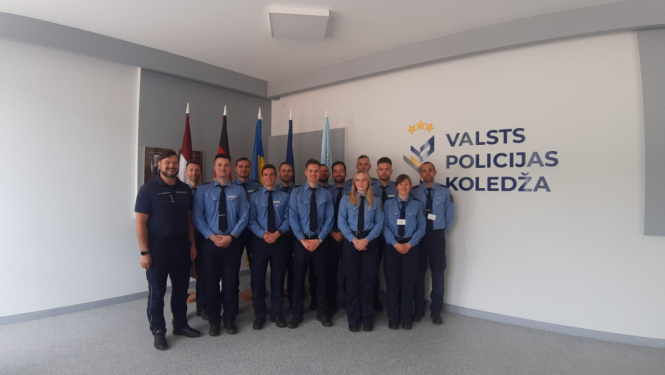 Grupas foto Valsts policijas koledžas telpās – formas tērpos sapulcējušies kadeti un instruktori stāv rindā pie sienas ar koledžas logotipu un karogiem fonā.