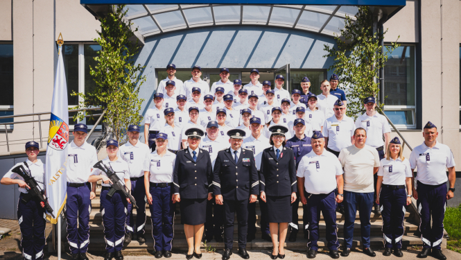 Grupas fotogrāfija pie Valsts policijas koledžas ēkas, kurā redzami policijas formas tērpos tērpti kadeti un instruktori, kā arī daži civilpersonas. Fonā redzama ēkas ieeja ar uzrakstu 'Valsts policijas koledža' un karogs ar Latvijas Valsts policijas emblēmu priekšplānā.