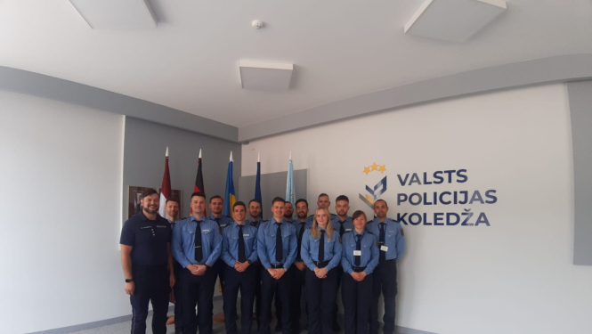 Grupas foto Valsts policijas koledžas telpās – formas tērpos sapulcējušies kadeti un instruktori stāv rindā pie sienas ar koledžas logotipu un karogiem fonā.