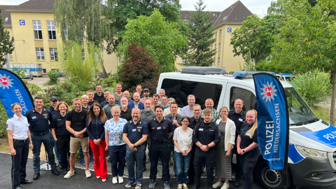 Grupas fotogrāfija, kurā redzami Vācijas Lejassaksijas policijas darbinieki un citi dalībnieki, stāvot kopā pie policijas busiņa ārpus ēkas ar uzrakstu 'Polizei Niedersachsen'. Fonā redzamas ēkas ar burtu 'C' un zaļojoši koki.