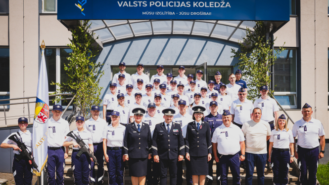 Grupas fotogrāfija pie Valsts policijas koledžas ēkas, kurā redzami policijas formas tērpos tērpti kadeti un instruktori, kā arī daži civilpersonas. Fonā redzama ēkas ieeja ar uzrakstu 'Valsts policijas koledža' un karogs ar Latvijas Valsts policijas emblēmu priekšplānā.