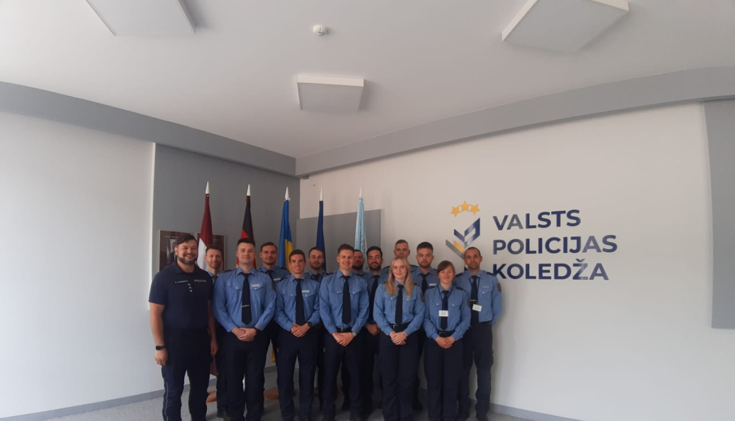 Grupas foto Valsts policijas koledžas telpās – formas tērpos sapulcējušies kadeti un instruktori stāv rindā pie sienas ar koledžas logotipu un karogiem fonā.
