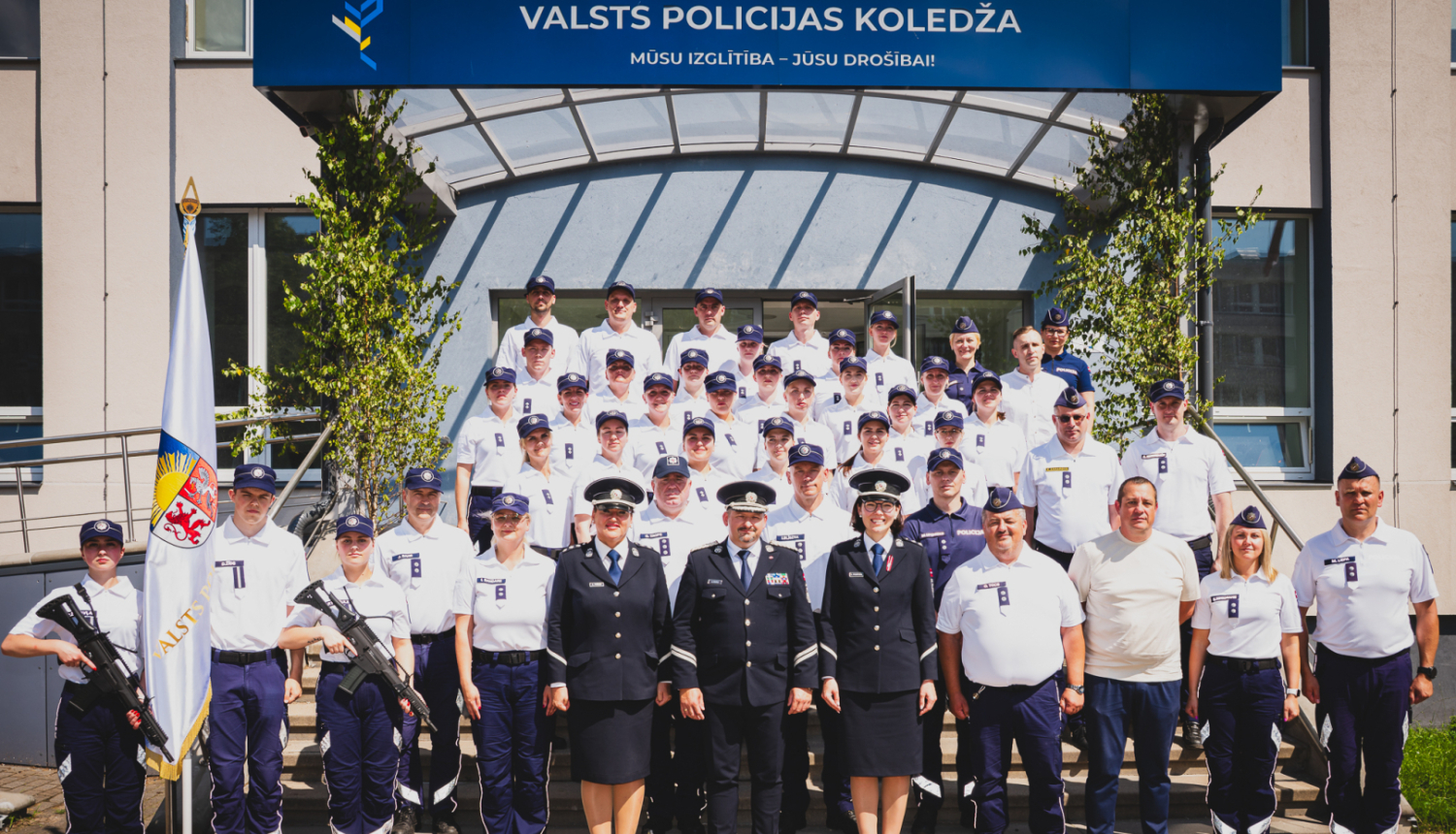 Grupas fotogrāfija pie Valsts policijas koledžas ēkas, kurā redzami policijas formas tērpos tērpti kadeti un instruktori, kā arī daži civilpersonas. Fonā redzama ēkas ieeja ar uzrakstu 'Valsts policijas koledža' un karogs ar Latvijas Valsts policijas emblēmu priekšplānā.