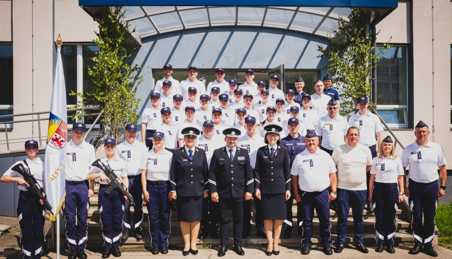 Grupas fotogrāfija pie Valsts policijas koledžas ēkas, kurā redzami policijas formas tērpos tērpti kadeti un instruktori, kā arī daži civilpersonas. Fonā redzama ēkas ieeja ar uzrakstu 'Valsts policijas koledža' un karogs ar Latvijas Valsts policijas emblēmu priekšplānā.
